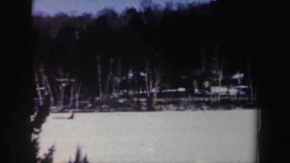 1962 New York home-movie frame showing a snow-covered lake, shadowed shoreline trees, evergreens, and a wooded hillside.