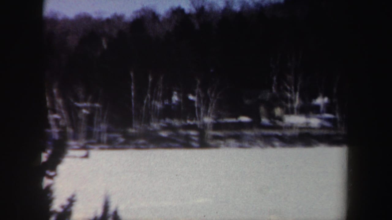 1962 New York 8mm frame showing a snowy frozen surface with leafless trees and evergreens along the far shore.