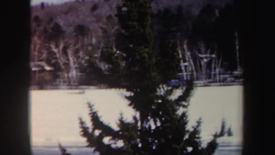 1962 New York winter frame showing a dark evergreen silhouette, snow-covered frozen surface, and bare shoreline trees.