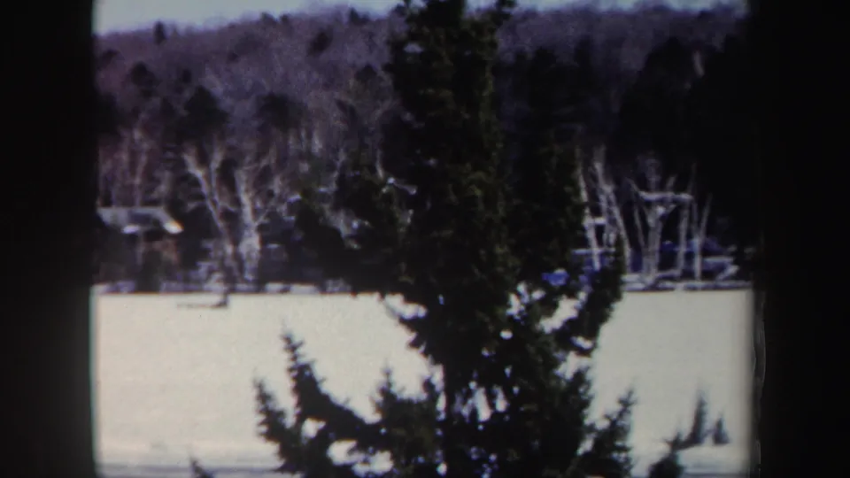 1962 New York winter landscape with a frozen lake or snowy open surface, dark evergreens, bare shoreline trees, and a small distant cabin-like structure.