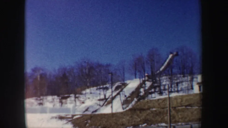1962 New York 8mm frame showing a snowy hillside, ski jump framework, bare trees, and clear blue winter sky.