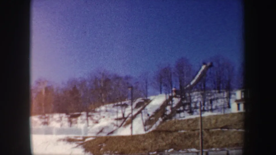 1962 New York winter frame with a ski jump structure on a snowy slope, bare trees along the ridge, and color-faded 8mm exposure.