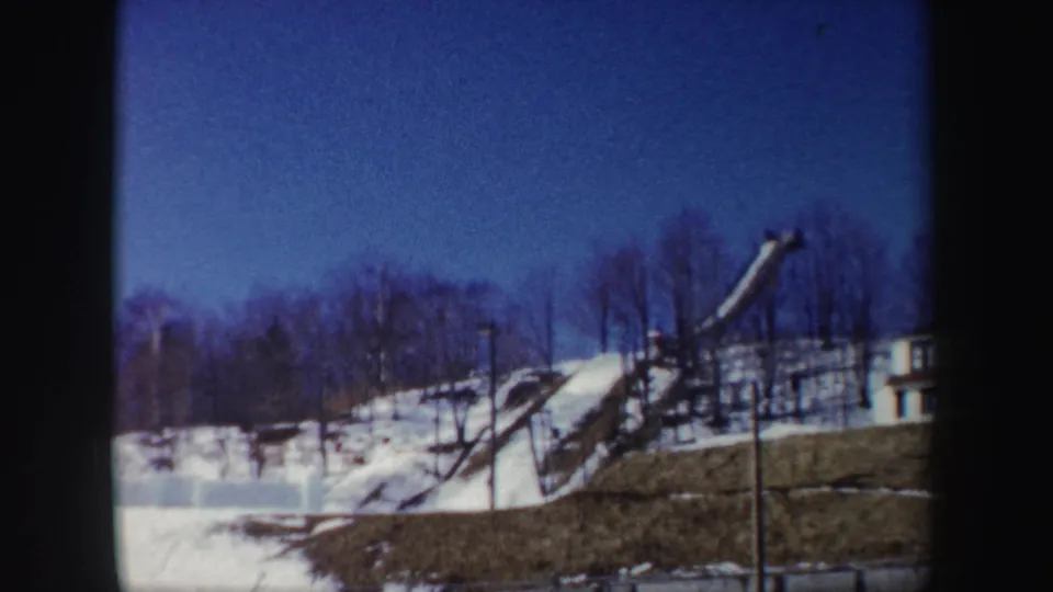 1962 New York home-movie frame of a snow-lined hill with bare trees, pole lights, and a curved elevated structure.