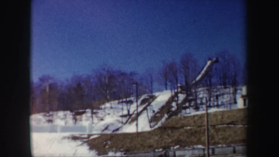1962 New York 8mm frame holding on the same snowy hillside, grassy foreground, bare trees, and possible recreational structure.