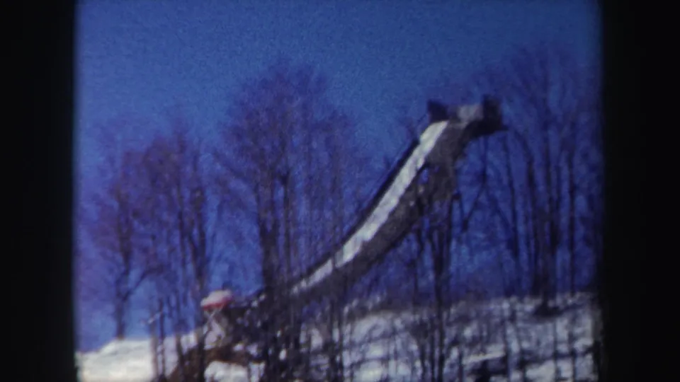 1962 New York 8mm frame with a long elevated structure crossing a winter hillside under a saturated blue sky.