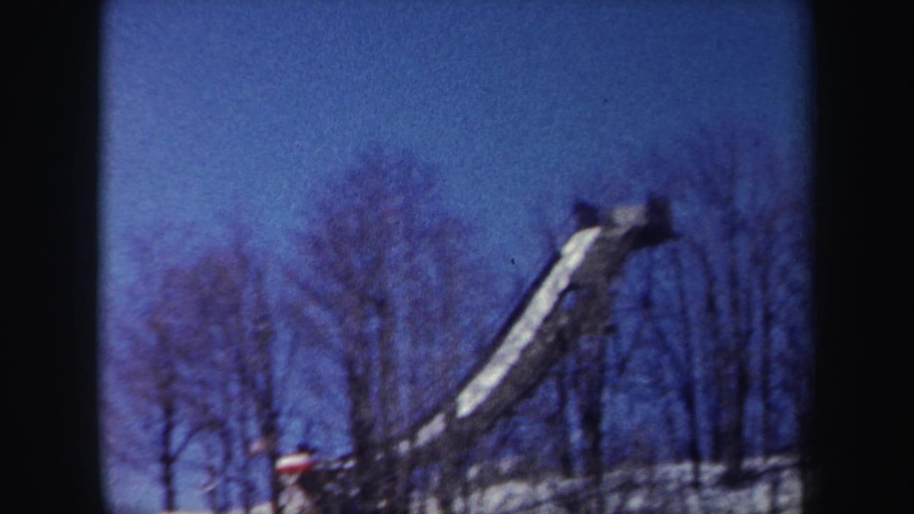 1962 New York 8mm frame showing a snow-covered wooded slope with bare trees and a long icy ramp or slide-like structure.