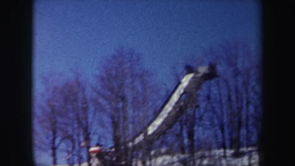 1962 New York 8mm frame showing a snow-covered wooded slope with bare trees and a long icy ramp or slide-like structure.