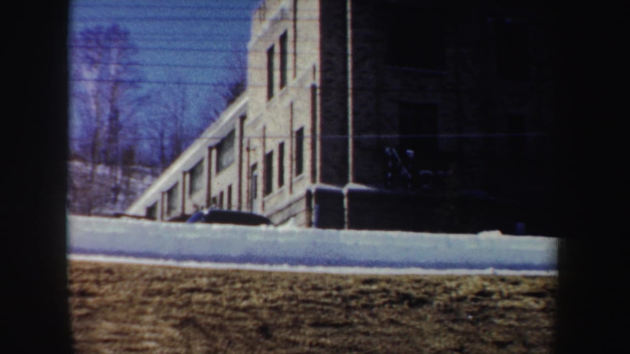 1962 New York 8mm frame showing a brick building corner, snow along the curb, bare trees, and winter sunlight.
