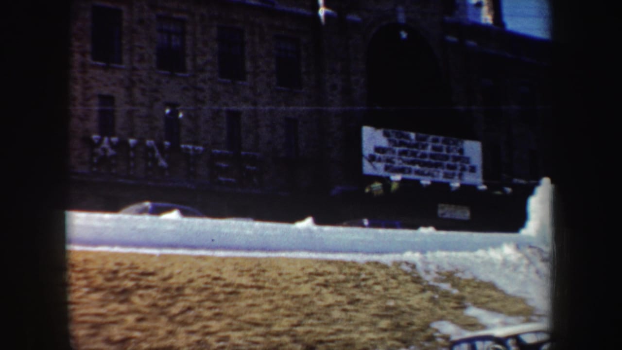 1962 New York 8mm frame showing snowbanks, a dark brick building facade, and a partially legible hanging event sign.