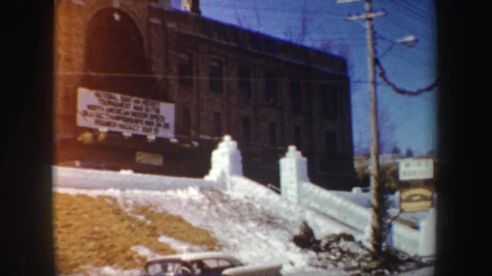 1962 New York home-movie frame with snowbanks, a brick building sign, utility pole, winter garland, and part of an older car.