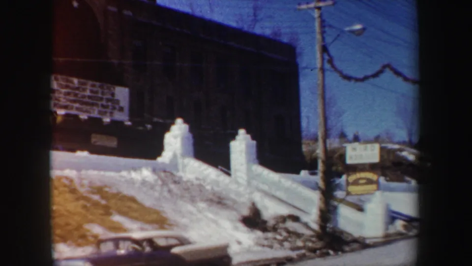 1962 New York winter street view showing the brick building sign, snow piles, utility pole, garland, and the roofline of a classic car.