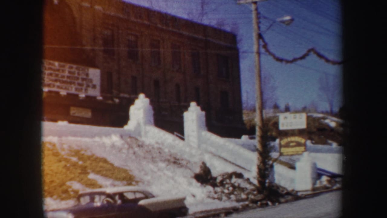1962 New York 8mm frame showing a snow-covered entrance, white gate posts, brick building, signboard, and part of a parked car.