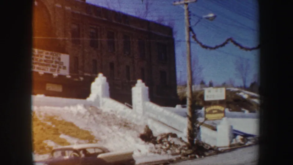 1962 New York home-movie view of a snowy gate outside a brick building, with a utility pole, signboard, and winter grounds.
