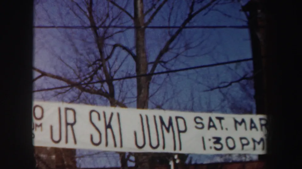 1962 New York frame with a hand-painted junior ski jump banner advertising a Saturday event at 1:30 PM below bare trees.