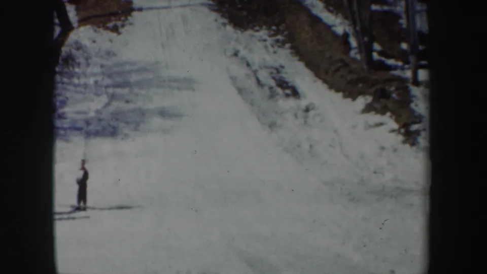 1962 New York 8mm frame showing a snow-covered ski-jump slope or run with one distant person standing below trees and shadowed snowbanks.