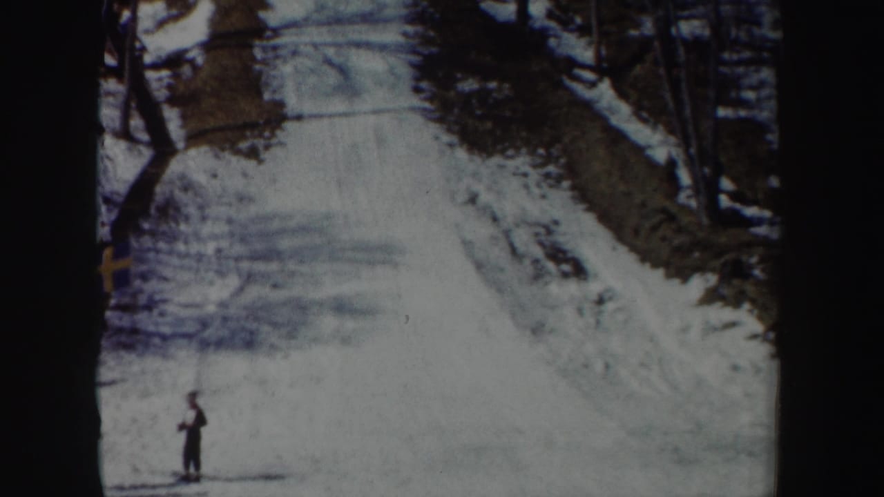 Snowy uphill ski-jump approach with bare trees and a small standing figure at lower left.