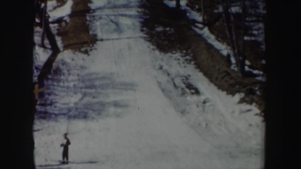 1962 New York 8mm frame showing a snow-covered ski-jump approach, bare trees, and a lone dark-clothed figure near the base.