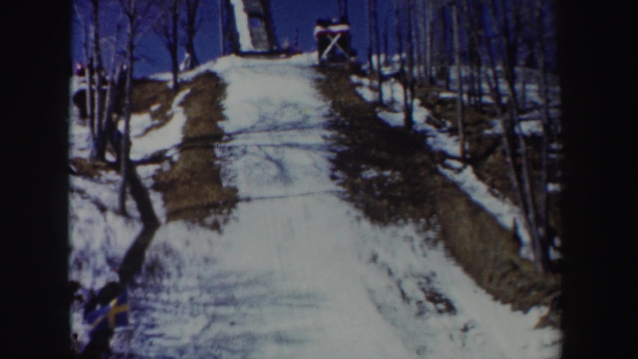 Snow-covered hill rising toward a wooden ski jump with bare trees and a small flag nearby.