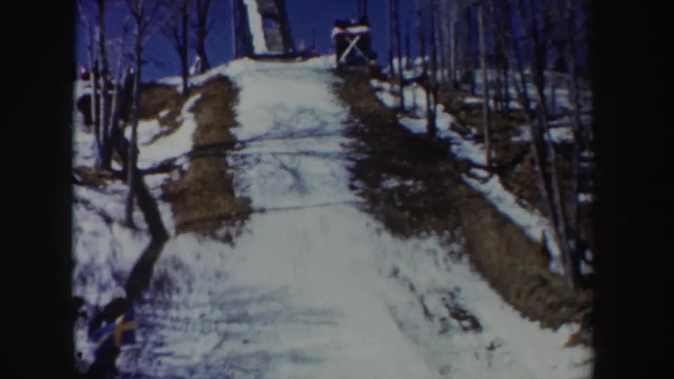 1962 New York 8mm frame looking up a snowy hill toward a wooden ski jump, with bare trees and a small flag at the side.