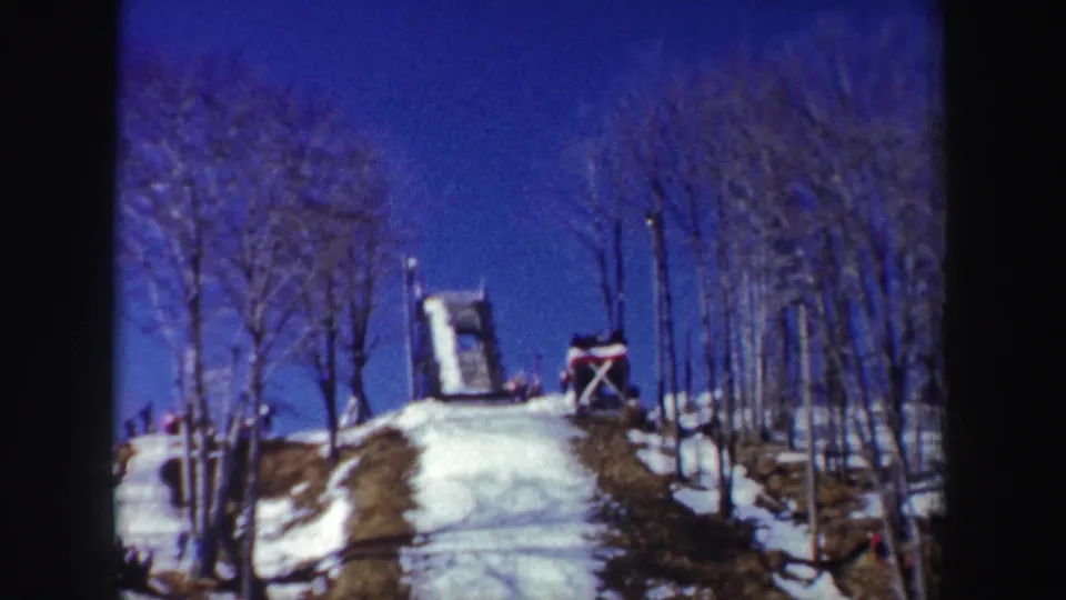 1962 New York 8mm frame showing the ski jump at the crest of a snowy wooded hill under clear blue sky.