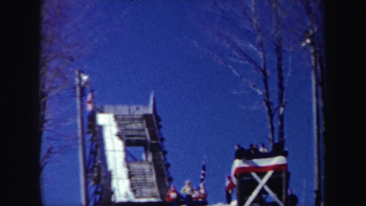 Wide view of a snowy ski jump ramp with bare trees, blue sky, and spectators near the base.