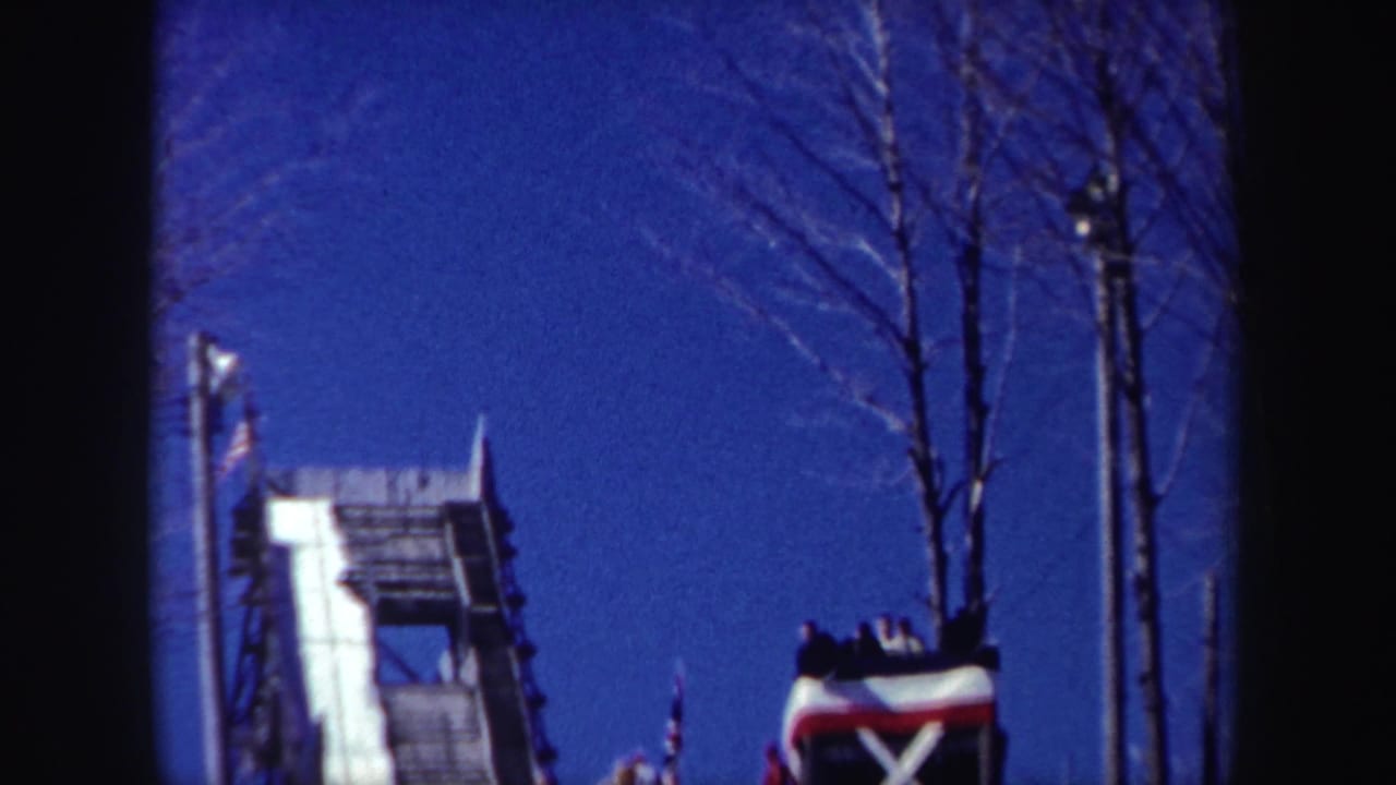 1962 New York home-movie frame with a snowy ski jump ramp, leafless trees, support poles, and small spectator groups.