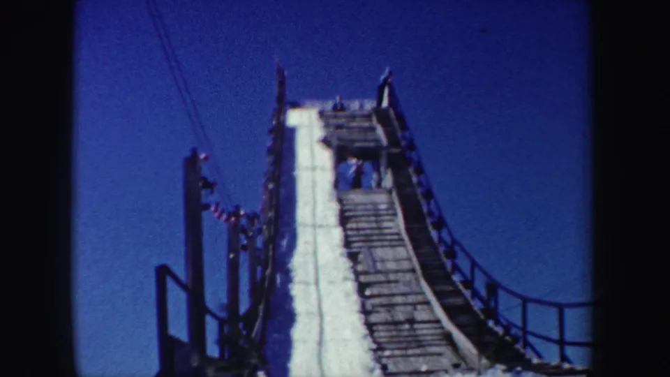 1962 New York frame showing the snow-covered ski jump ramp in closer detail, with wood construction, railings, and a clear blue sky.
