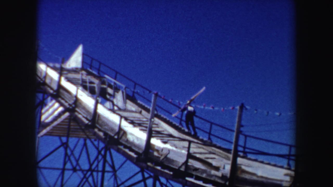 Worker carrying a long wooden piece up a steep railed structure under blue sky.
