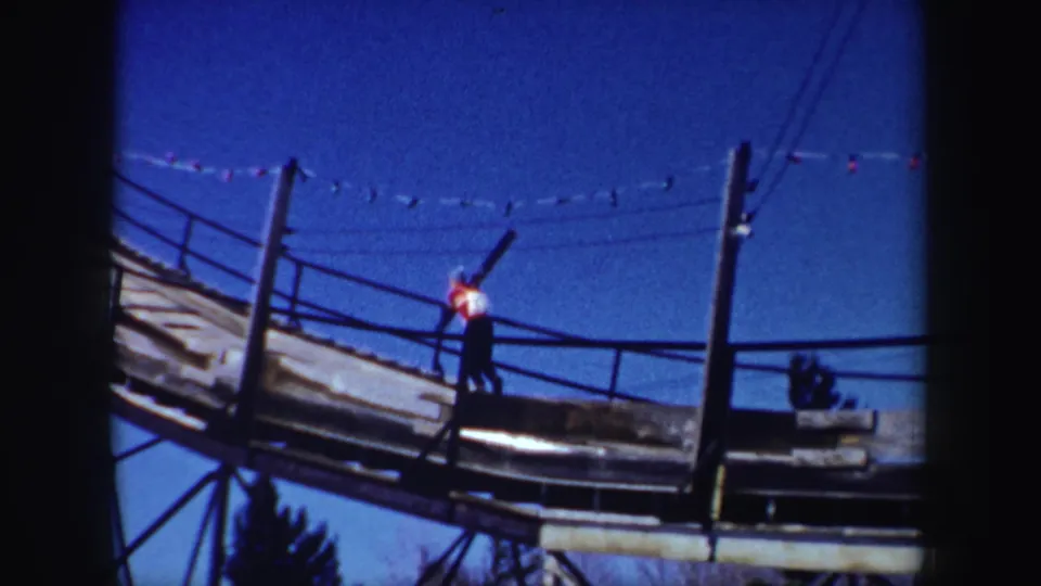 1962 New York 8mm frame with a worker on an elevated railed structure, seen beneath a clear blue sky.