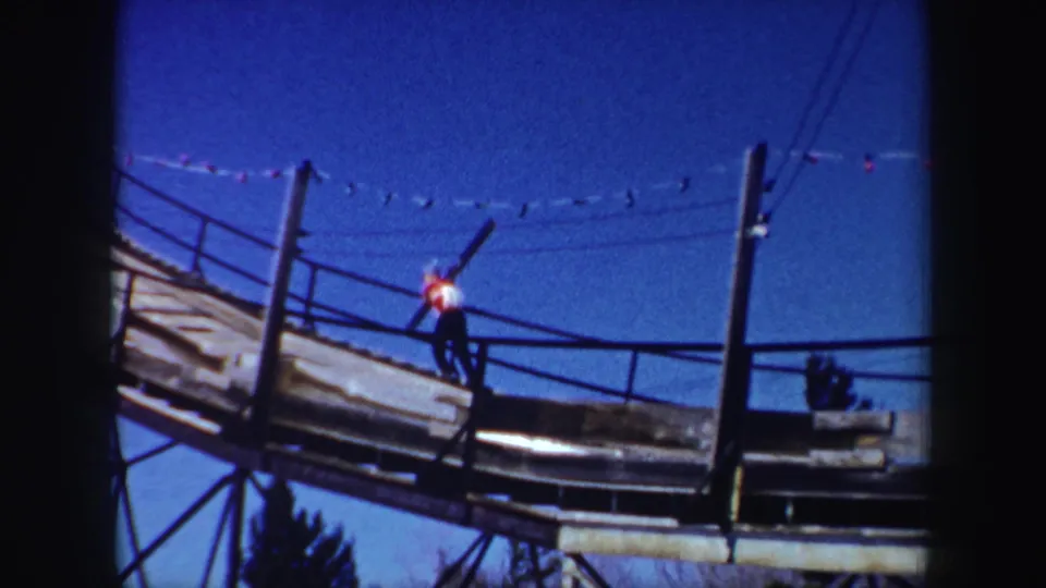 In 1962 New York home-movie footage, a skier in a red-and-white jacket carries skis up a wooden ramp beneath overhead lights.