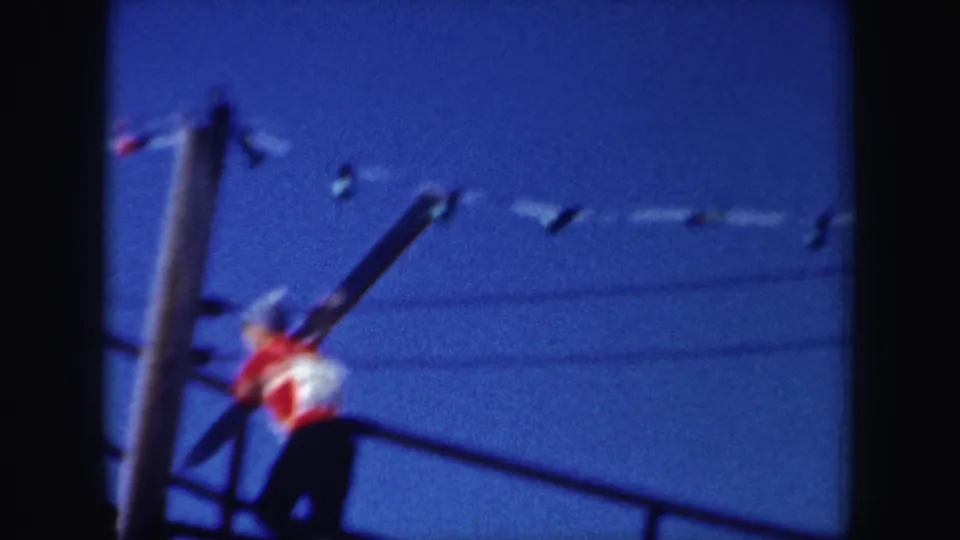 The skier appears partly obscured and motion-blurred while carrying skis beside a pole on the ramp, with lights crossing the sky above.