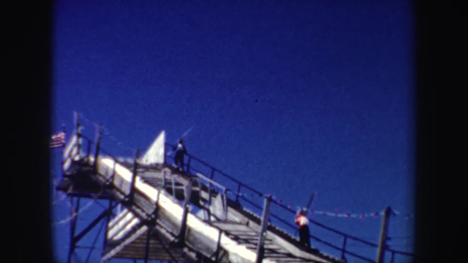 1962 New York 8mm frame showing a ski-area stairway with two people on it, pennant flags, and an American flag at the upper platform.