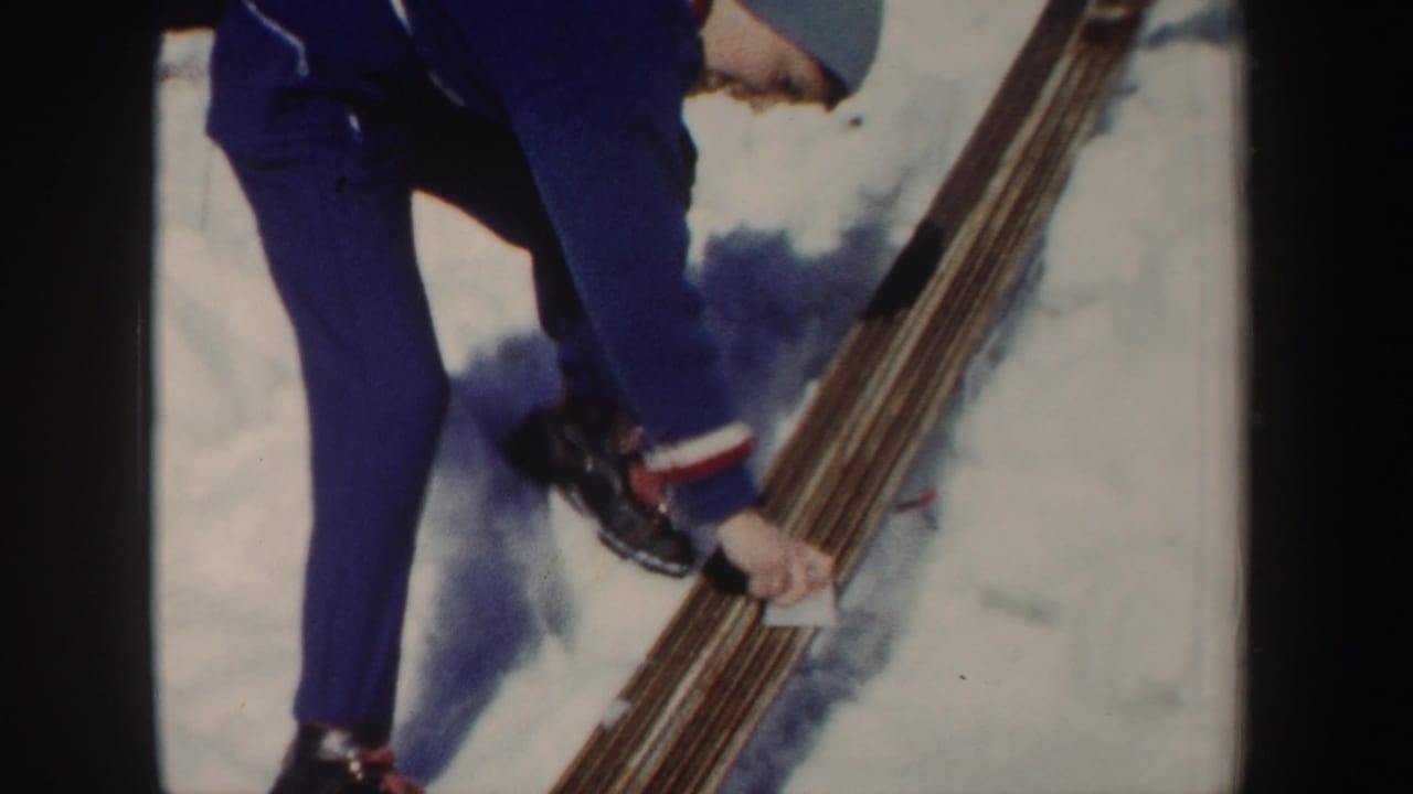 Skier's lower body and hands near a bundle of wooden skis on snow.