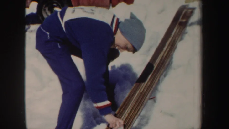 1962 New York 8mm frame of a skier in a blue outfit and gray cap bending to prepare or clean wooden skis.