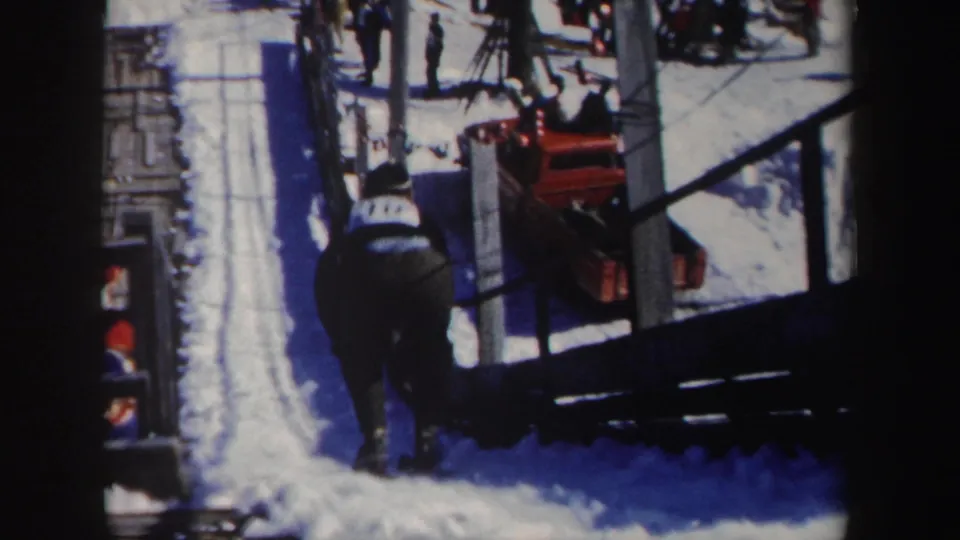 A skier approaches or passes along a snow-covered course in the 1962 New York footage, with spectators, fencing, bare trees, and a red vehicle visible.