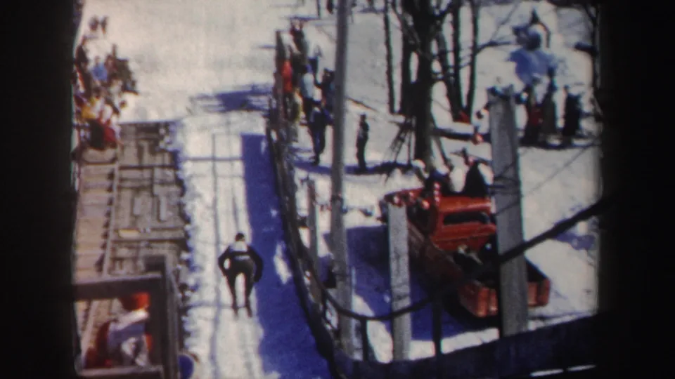 The ski course is seen from above or the side, with a skier on the snow, spectators along the edge, fencing, trees, and a red vehicle in frame.