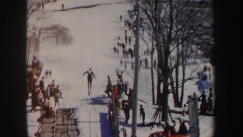 Wide 8mm view of the snowy ski-jump area in New York, with a skier in the landing zone, spectators along the sidelines, and bare trees nearby.