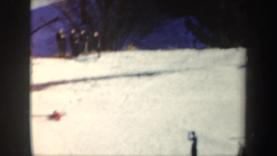 1962 New York home-movie frame of a red-clad skier mid-jump on a sunny snow slope with distant onlookers.