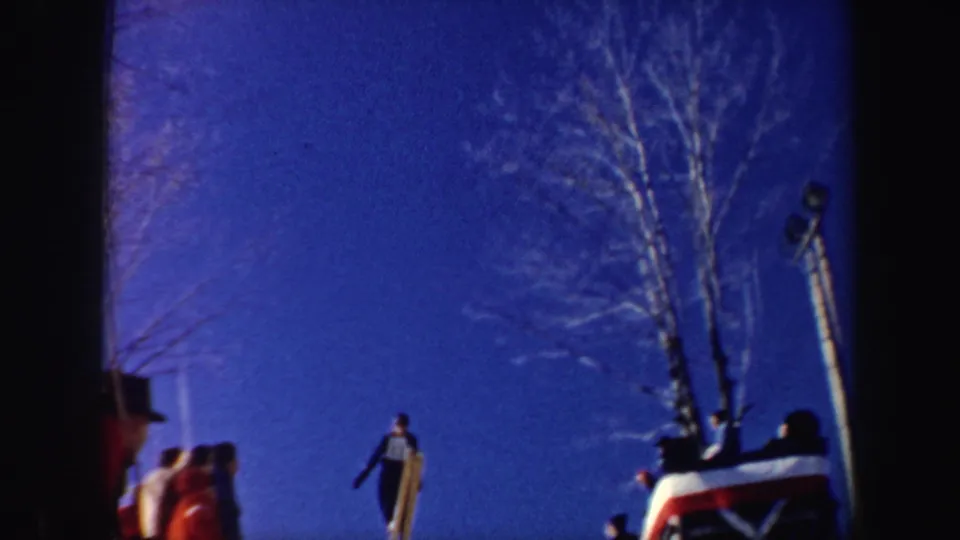 1962 New York 8mm frame showing a skier in a dark outfit descending the slope with spectators and bare trees in the background.