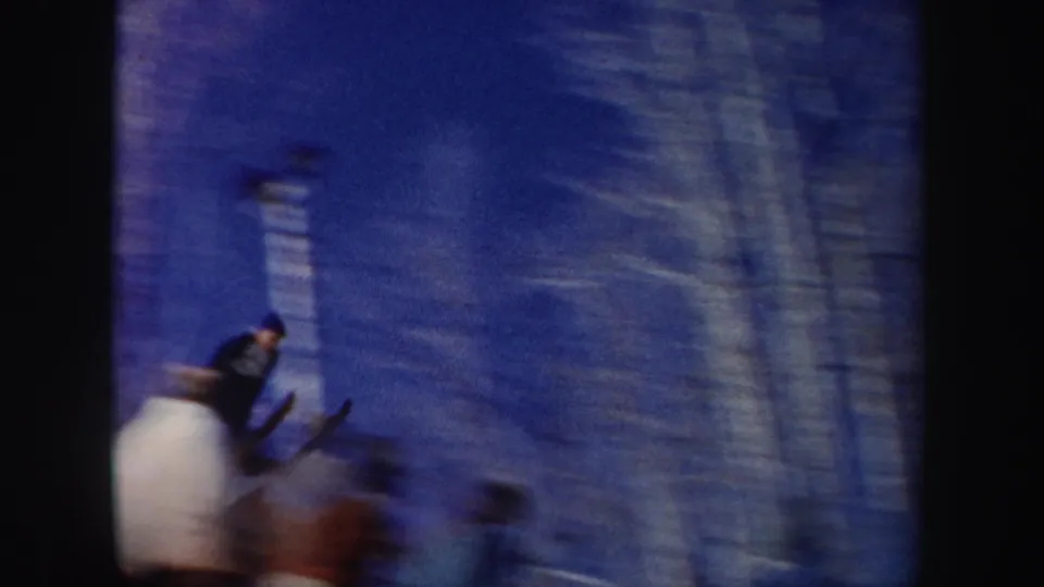 A skier in blue appears airborne or descending on a snowy New York slope, with blurred onlookers near the lower frame.