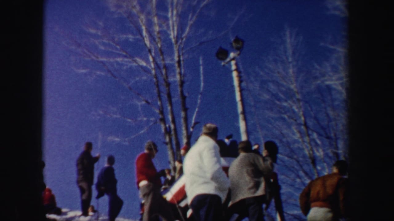 1962 New York home-movie frame of a bundled crowd watching from a snowy hillside beside bare trees and a light pole.