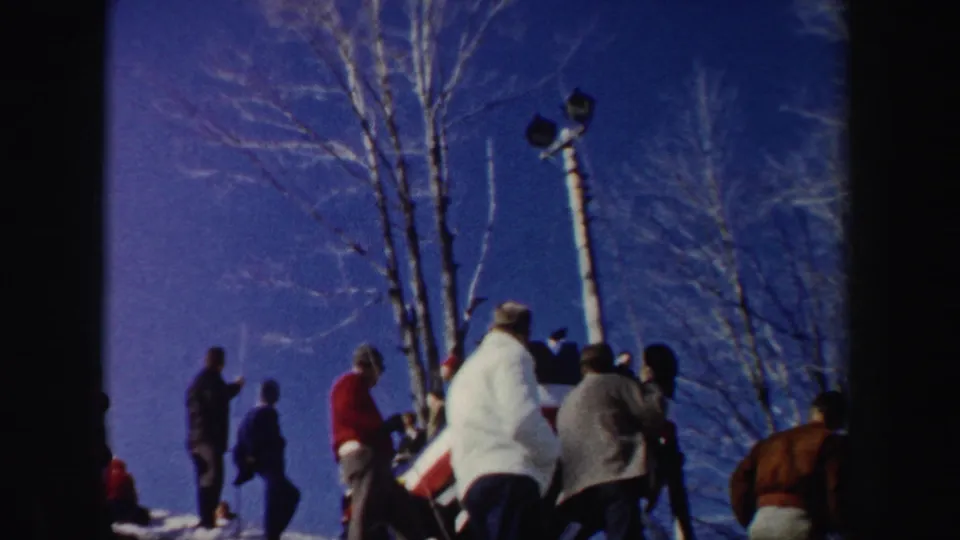 1962 New York home-movie frame of a bundled crowd watching from a snowy hillside beside bare trees and a light pole.
