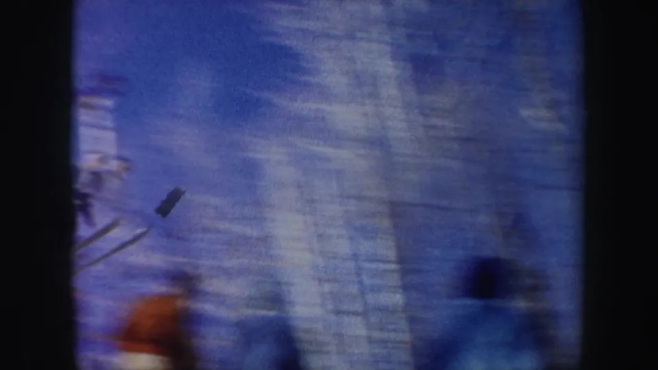 A distant ski jumper is captured midair in a soft, handheld 8mm frame with snow, blue sky, and blurred foreground movement.