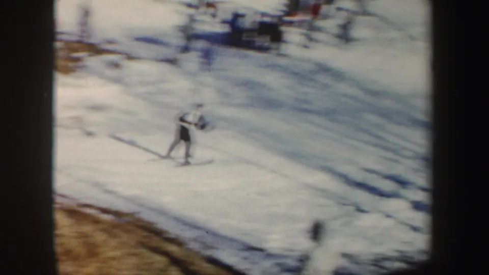 Wide home-movie view of a skier on a snowy hillside, with ski tracks and distant spectators or equipment near the top of frame.