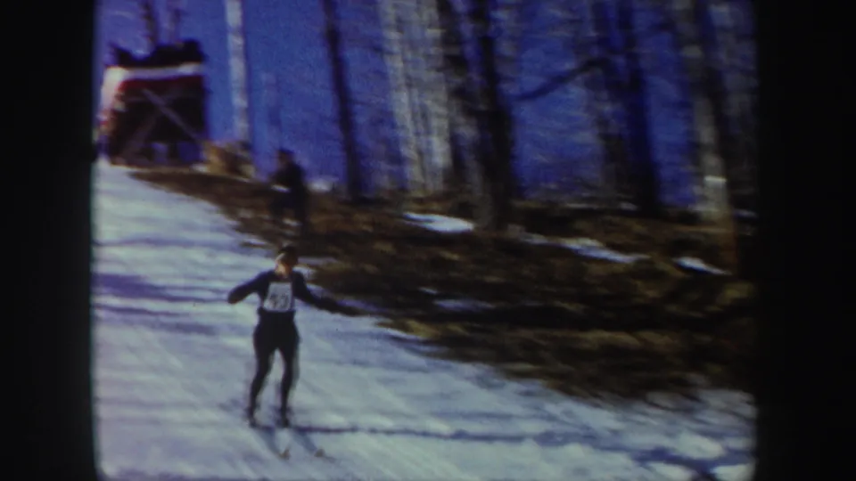 1962 New York home-movie frame of a skier in a numbered bib moving downhill on snow, with bare trees and a small structure near the top.