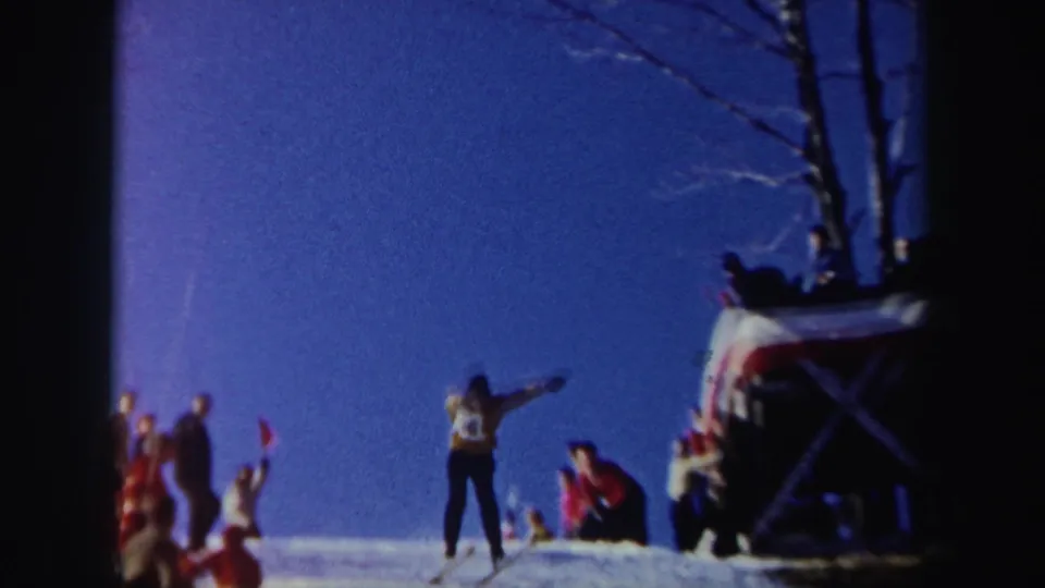 1962 New York 8mm frame showing spectators near a small ramp area while a skier in a numbered bib stands or moves on the slope.