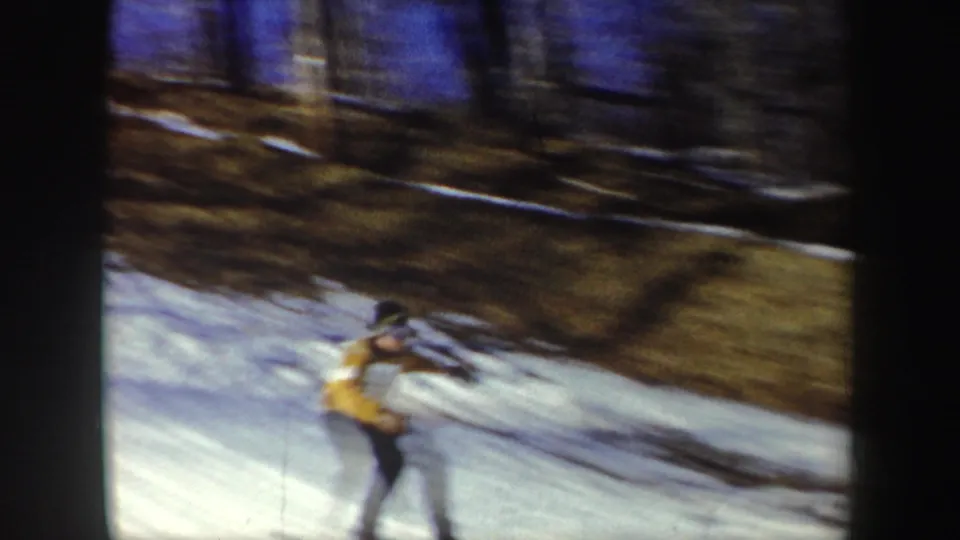 1962 New York home-movie frame of a skier in a yellow jacket descending across a snowy slope, blurred by motion and handheld filming.