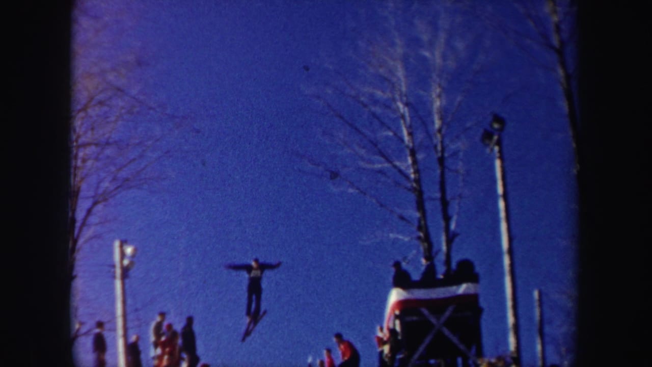 In 1962 New York 8mm footage, a skier in dark clothing is airborne near a jump structure as spectators stand on the snowy slope.