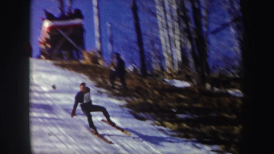 The skier lands or descends on the snowy New York slope, with bare trees and the jump structure visible behind him.