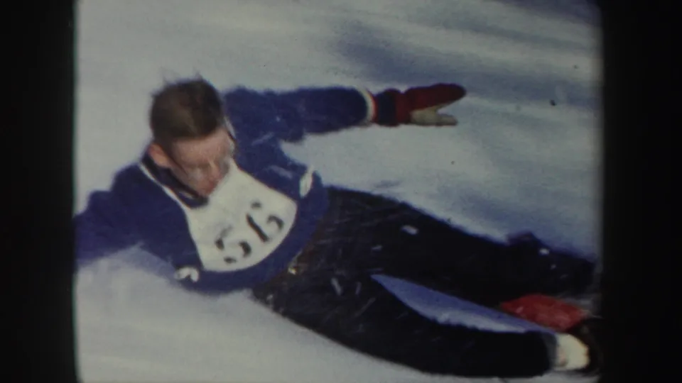 A skier wearing a blue outfit and white numbered bib slides low across the snow after the landing in this 1962 home-movie frame.
